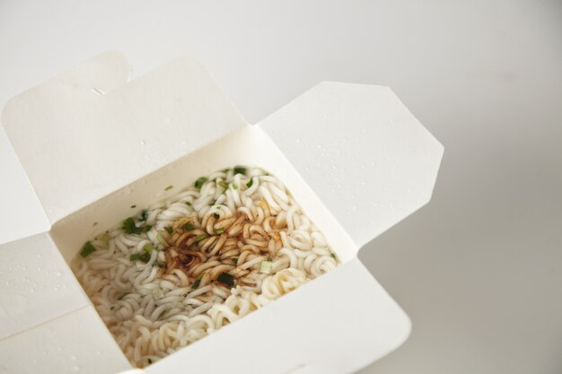 Vista superior de cerca en la sabrosa sopa de fideos con pasta y salsa de soja dentro de la caja de papel para llevar en blanco aislado en blanco