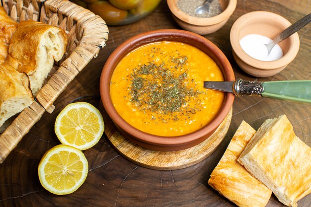 Una vista superior de cerca sopa de merji naranja dentro de una olla marrón junto con rodajas de limones y rebanadas de pan