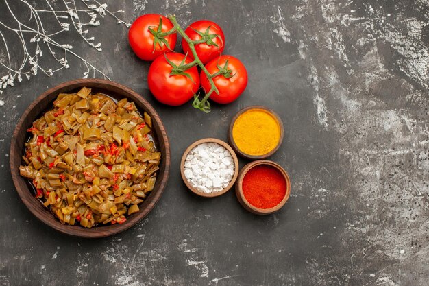 Vista superior de cerca tazones de especias de especias y tomates junto al plato de judías verdes y tomates con pedicelo en la mesa oscura