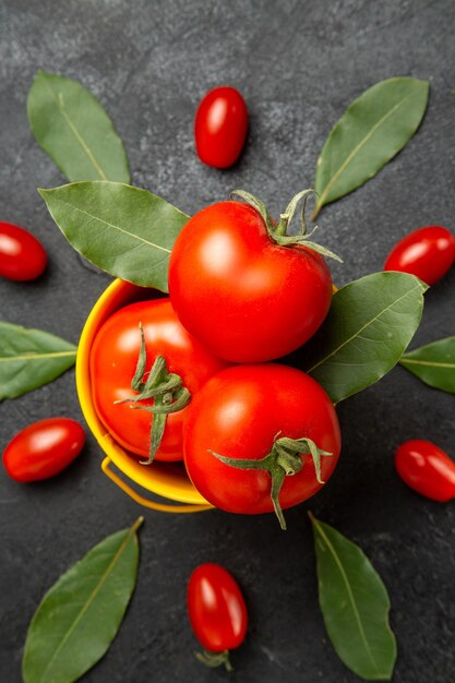 Vista superior cercana de un balde con tomates alrededor de tomates cherry y hojas de laurel sobre suelo oscuro