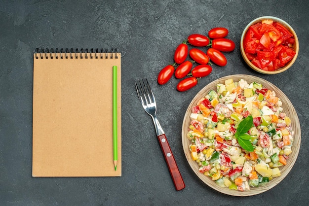 Vista superior cercana de ensalada de verduras con tenedor de tomates y bloc de notas en el lado sobre fondo gris oscuro