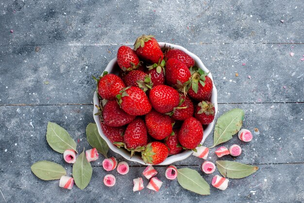 Vista superior cercana de fresas rojas frescas dentro de la placa junto con caramelos rosados en rodajas en gris, fruta baya color foto fresca suave