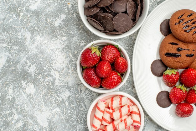 Vista superior cercana galletas de chocolate fresas y chocolates redondos en la placa ovalada blanca y cuencos con dulces fresas chocolates en el suelo gris-blanco