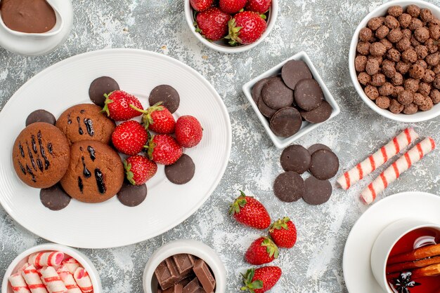 Vista superior cercana galletas fresas y bombones redondos en la placa ovalada cuencos de dulces fresas chocolates cereales cacao y té de canela en la mesa gris-blanca