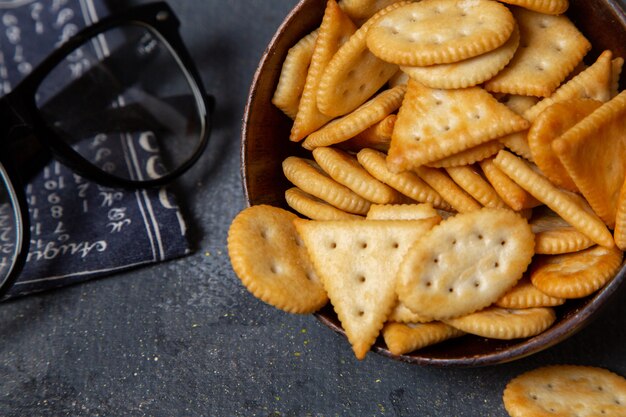 Vista superior cercana galletas saladas sabrosas dentro de un plato marrón con gafas de sol en el escritorio gris bocadillo de galletas crujientes