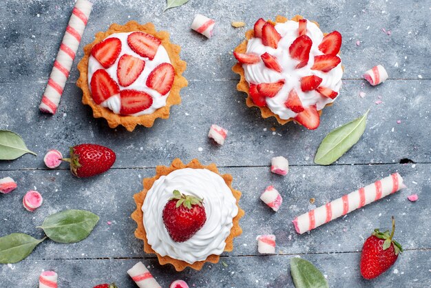 Vista superior cercana de pequeños pasteles cremosos con fresas frescas y en rodajas junto con caramelos de palo en gris