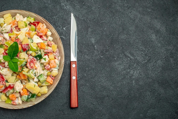Vista superior cercana del tazón de fuente de ensalada de verduras con cuchillo sobre fondo gris oscuro