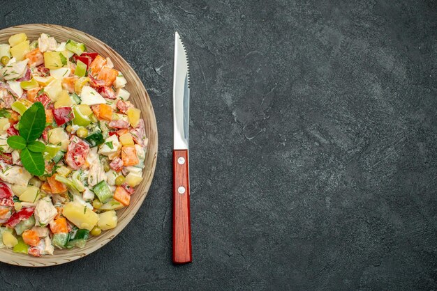 Vista superior cercana del tazón de fuente de ensalada de verduras con cuchillo sobre fondo gris oscuro