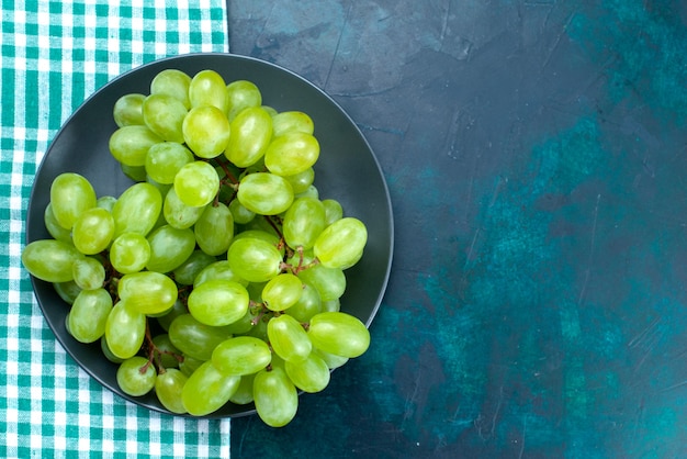 Vista superior cercana uvas verdes frescas frutas jugosas suaves dentro de la placa en el escritorio azul oscuro.