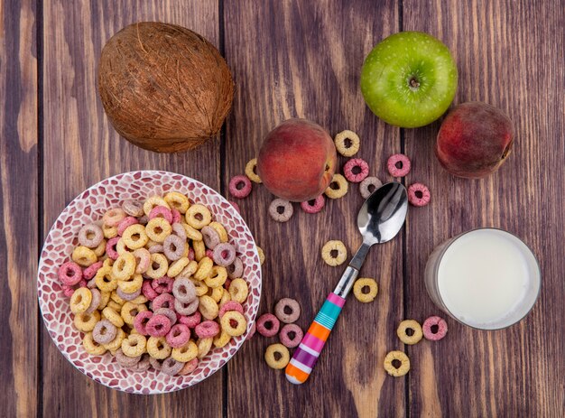 Vista superior de cereales en un tazón con una cuchara con frutas frescas como manzana, melocotón y un vaso de leche en madera