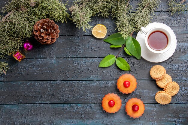 Vista superior de la cereza cupcakes cono ramas de abeto rodaja de limón una taza de té y galletas en el suelo de madera oscura con espacio de copia