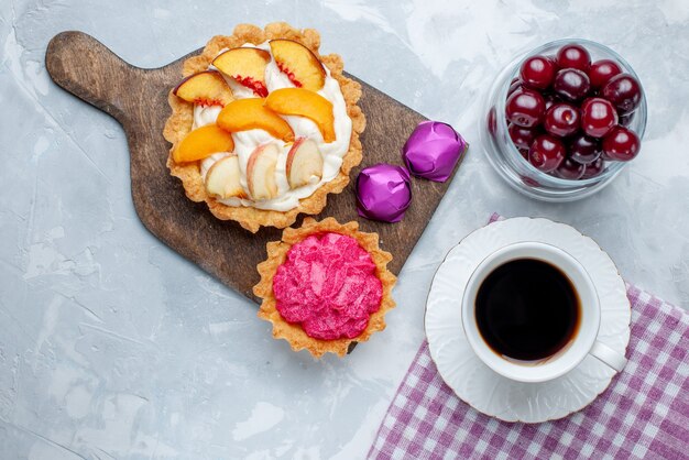 Vista superior de cerezas ácidas frescas dentro de un pequeño vaso de vidrio con tortas de crema y té en el escritorio de luz blanca, fruta agria vitamina dulce