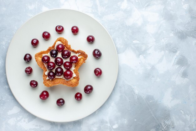 Vista superior de las cerezas ácidas frescas dentro de la placa con torta cremosa en forma de estrella en blanco claro, frutas, bayas ácidas, vitamina verano