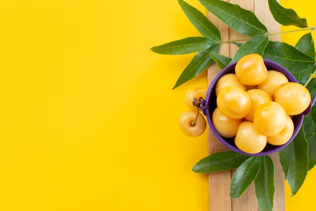 Una vista superior de las cerezas amarillas suaves y dulces con hojas verdes en el escritorio amarillo, color de la fruta cereza dulce de verano