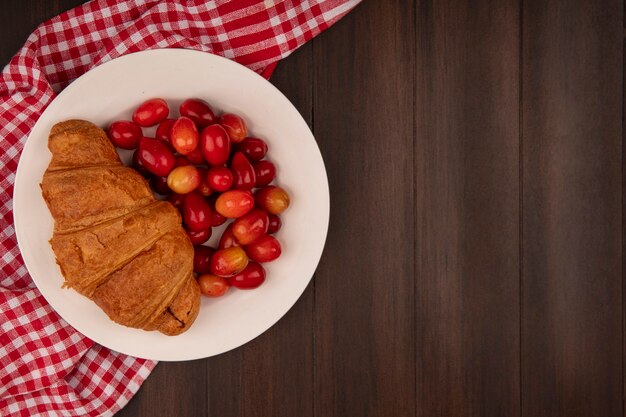 Vista superior de las cerezas de cornalina agria roja con croissant en un plato blanco sobre un paño rojo marcado en una pared de madera con espacio de copia