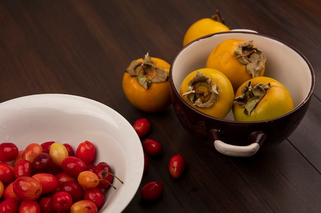 Foto gratuita vista superior de las cerezas de cornalina en un recipiente blanco con frutos de caqui amarillo en un recipiente sobre una superficie de madera