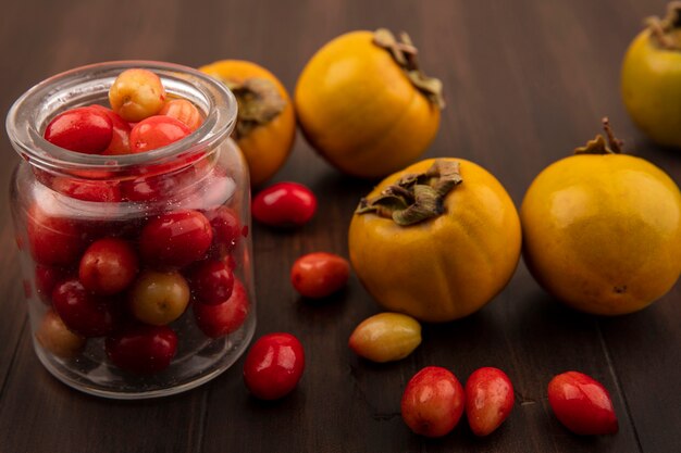 Vista superior de cerezas de cornalina roja en un frasco de vidrio con frutos de caqui aislado en una superficie de madera