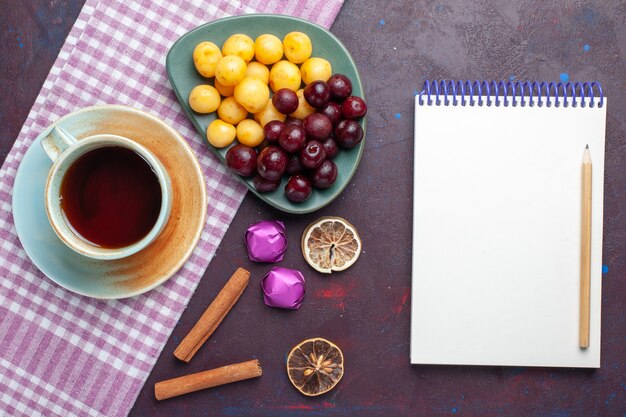 Vista superior de cerezas dulces frescas dentro de la placa con una taza de té y un bloc de notas en la superficie oscura