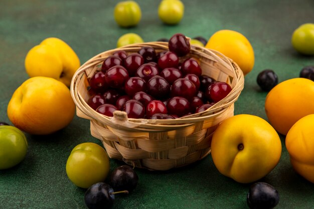 Vista superior de cerezas rojas en un cubo con melocotones amarillos con ciruelas cereza verde sobre un fondo verde