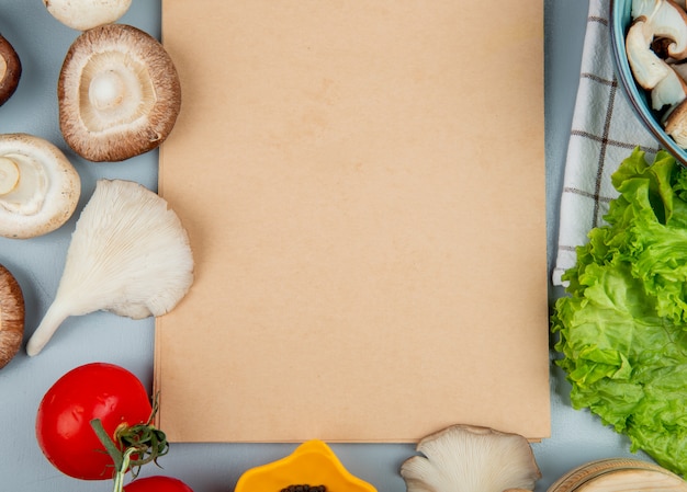 Vista superior de champiñones frescos con tomate lechuga dispuestos alrededor de un cuaderno de bocetos en azul claro