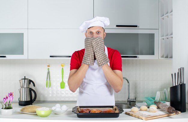 Vista superior del chef asustado con soporte de pie detrás de la mesa con pasteles rallador de huevos en la cocina blanca