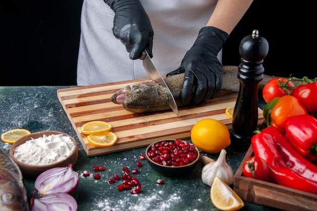 Vista superior del chef cortando la cabeza de pescado en la tabla de cortar, molinillo de pimienta, tazón de harina, semillas de granada en un tazón, verduras en la mesa de la cocina