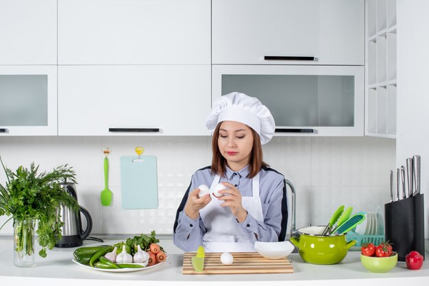 Vista superior del chef enfocado y verduras frescas con equipo de cocina y sosteniendo huevos en la cocina blanca