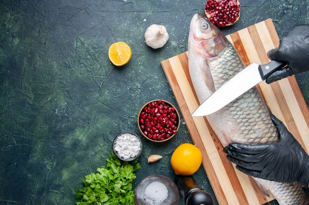 Vista superior del chef con guantes negros cortando pescado crudo en la tabla de cortar semillas de granada molinillo de pimienta en un tazón en el lugar de la copia de la mesa de la cocina