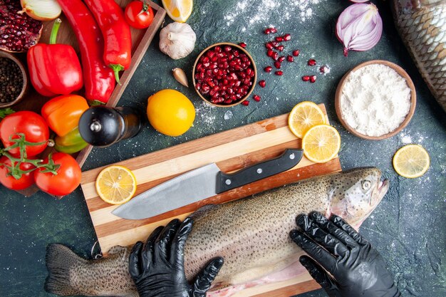 Vista superior del chef con guantes negros sosteniendo pescado crudo en tablero de madera molinillo de pimienta harina tazón de granada semillas en un tazón en la mesa de la cocina