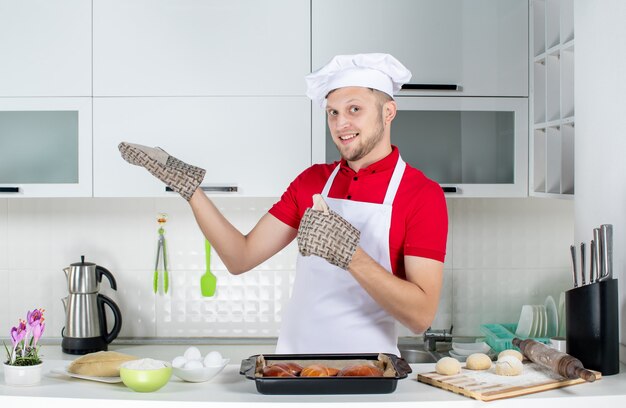 Vista superior del chef masculino confiado con un soporte de pie detrás de la mesa con un rallador de huevos de pasteles y mostrando algo en el lado derecho en la cocina blanca