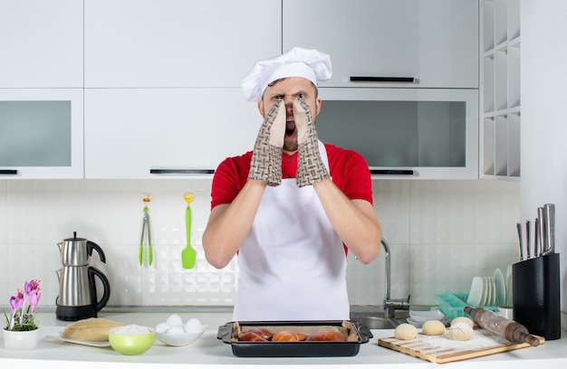 Vista superior del chef masculino con soporte de pie detrás de la mesa con pasteles rallador de huevos y llamando a alguien en la cocina blanca