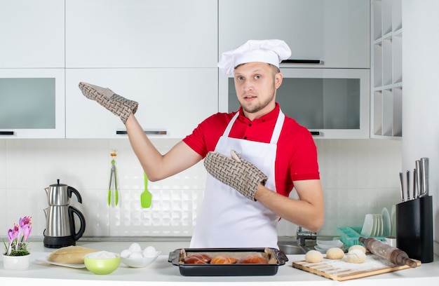 Vista superior del chef masculino con soporte de pie detrás de la mesa con pasteles rallador de huevos y mostrando algo en el lado derecho en la cocina blanca