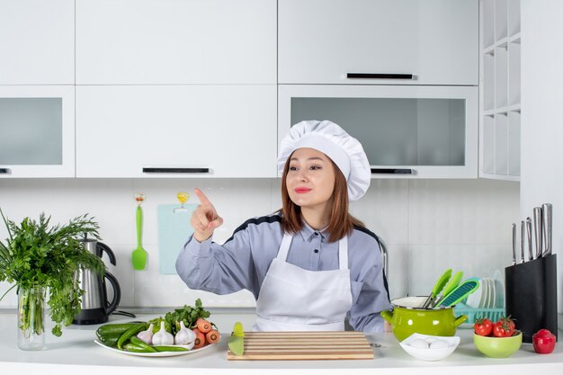 Vista superior de la chef mujer sonriente y verduras frescas con equipo de cocina y apuntando hacia adelante en la cocina blanca