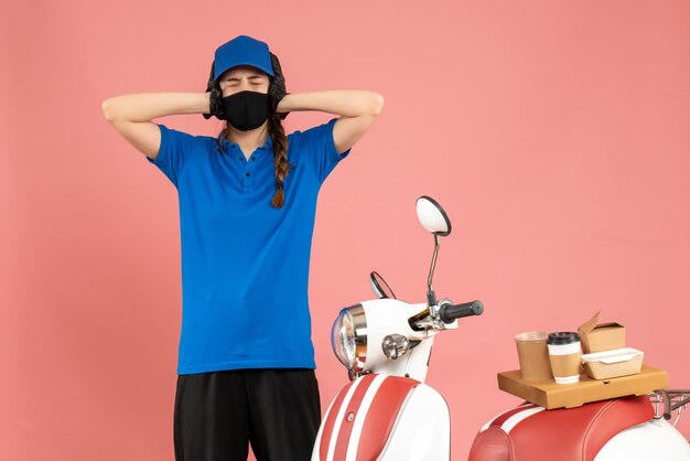 Vista superior de la chica de mensajería agotada en máscara médica de pie junto a la motocicleta con pastel de café sobre fondo de color melocotón pastel