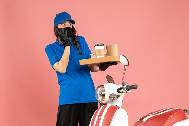 Vista superior de la chica de mensajería asustada con guantes de máscara médica de pie junto a la motocicleta con pequeños pasteles de café sobre fondo de color melocotón pastel