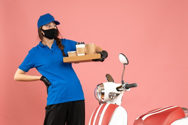 Vista superior de la chica de mensajería asustada con guantes de máscara médica de pie junto a la motocicleta sosteniendo pequeños pasteles de café sobre fondo de color melocotón pastel