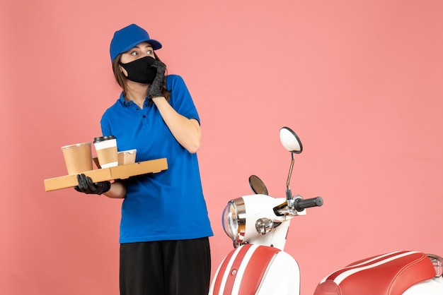 Vista superior de la chica de mensajería asustada con guantes de máscara médica de pie junto a la motocicleta sosteniendo pequeños pasteles de café sobre fondo de color melocotón pastel