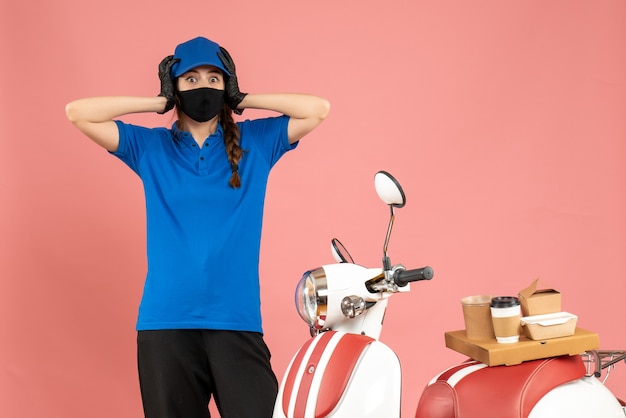 Vista superior de la chica de mensajería aterrorizada en máscara médica de pie junto a la motocicleta con pastel de café sobre fondo de color melocotón pastel