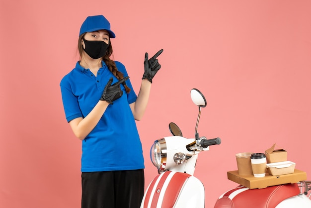 Vista superior de la chica de mensajería confiada con guantes de máscara médica de pie junto a la motocicleta con pastel de café apuntando hacia arriba sobre fondo de color melocotón pastel