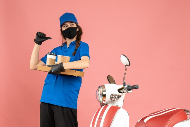 Vista superior de la chica de mensajería confiada con guantes de máscara médica de pie junto a la motocicleta sosteniendo pequeños pasteles de café apuntando hacia atrás sobre fondo de color melocotón pastel