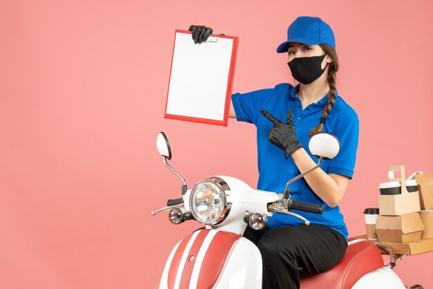 Vista superior de la chica de mensajería confiada con máscara médica y guantes sentado en scooter sosteniendo una hoja de papel vacía entregando pedidos sobre fondo melocotón pastel