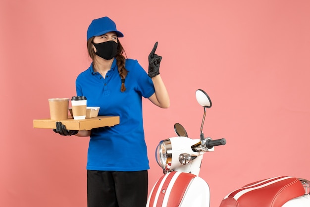 Vista superior de la chica de mensajería emocional con guantes de máscara médica de pie junto a la motocicleta sosteniendo pequeños pasteles de café apuntando hacia arriba sobre fondo de color melocotón pastel