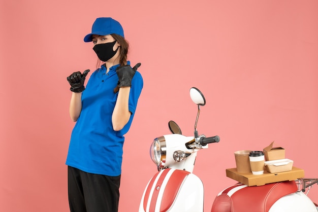 Vista superior de la chica de mensajería con guantes de máscara médica de pie junto a la motocicleta con pastel de café apuntando hacia atrás sobre fondo de color melocotón pastel