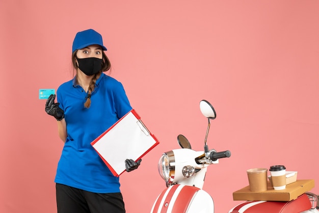 Vista superior de la chica de mensajería con guantes de máscara médica de pie junto a la motocicleta con pastel de café sosteniendo documentos tarjeta bancaria sobre fondo de color melocotón pastel