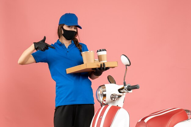 Vista superior de la chica de mensajería con guantes de máscara médica de pie junto a la motocicleta señalando pequeños pasteles de café sobre fondo de color melocotón pastel