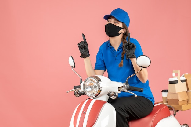 Vista superior de la chica de mensajería con máscara médica y guantes sentado en scooter entregando pedidos apuntando hacia arriba sobre fondo melocotón pastel