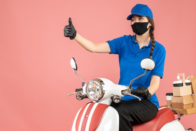 Vista superior de la chica de mensajería con máscara médica y guantes sentado en scooter entregando pedidos haciendo gesto ok sobre fondo melocotón pastel