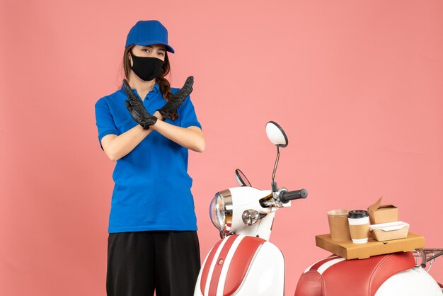 Vista superior de la chica de mensajería en máscara médica de pie junto a la motocicleta con pastel de café haciendo gesto de parada sobre fondo de color melocotón pastel