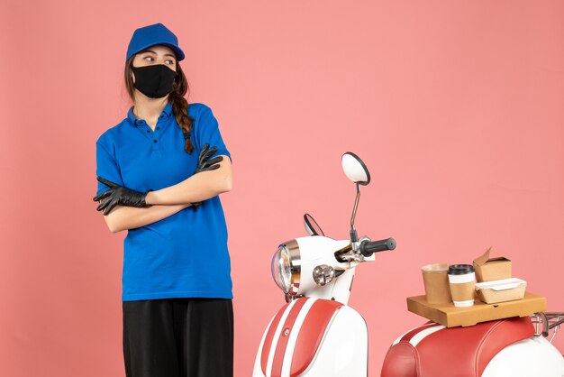 Vista superior de la chica de mensajería pensativa en máscara médica de pie junto a la motocicleta con pastel de café en color melocotón pastel