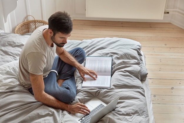 La vista superior de un chico barbudo relajado posa en una cama acogedora en posición de loto, reflexiona sobre el material leído, verifica la información del libro en la computadora portátil, estudia leyes, trabaja en el dormitorio. Atmósfera doméstica